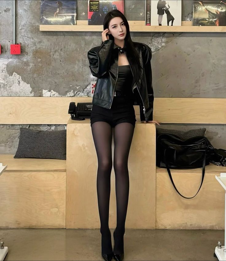 Woman in a black leather jacket, shorts, and pantyhose standing indoors