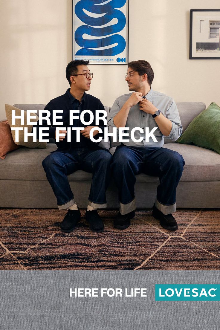 Two men sitting on a sofa in a living room, one adjusting his shirt