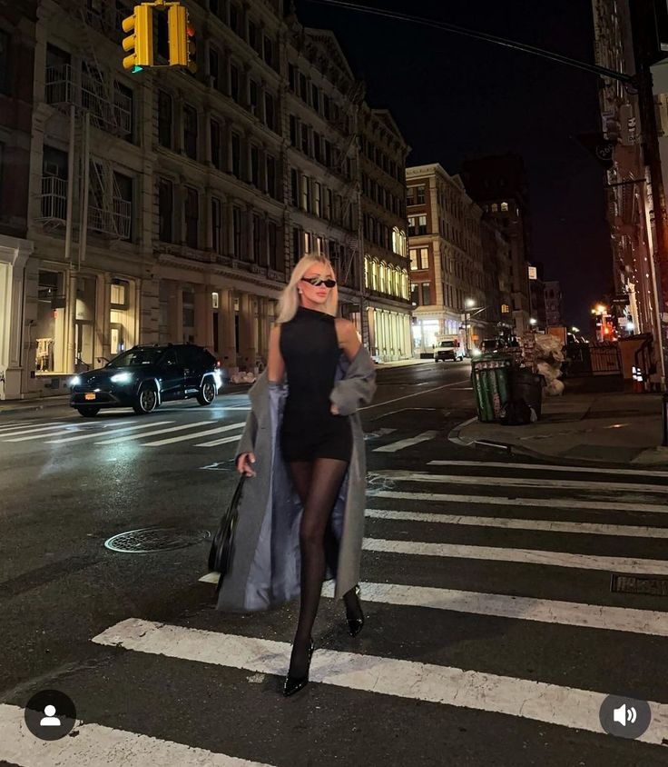 Woman in a black dress and long coat crossing a city street at night