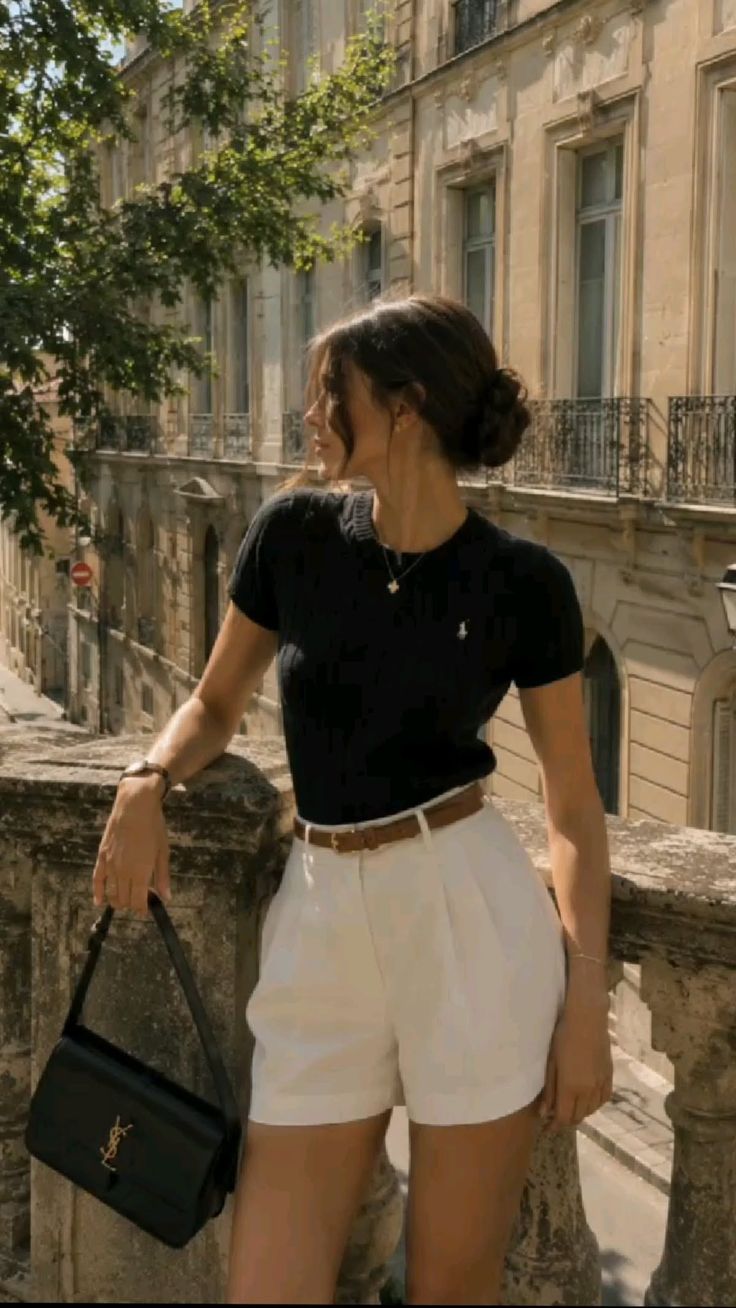 Woman in white shorts and a black top standing on a stone balcony overlooking a city