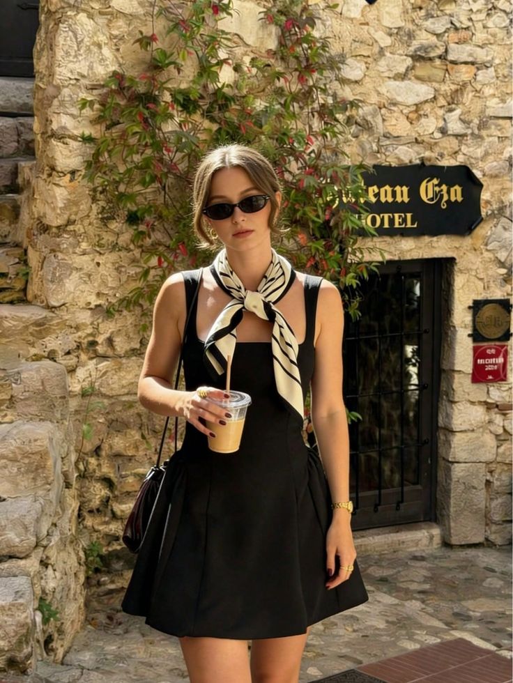 Woman in a black dress and sunglasses holding a drink outdoors