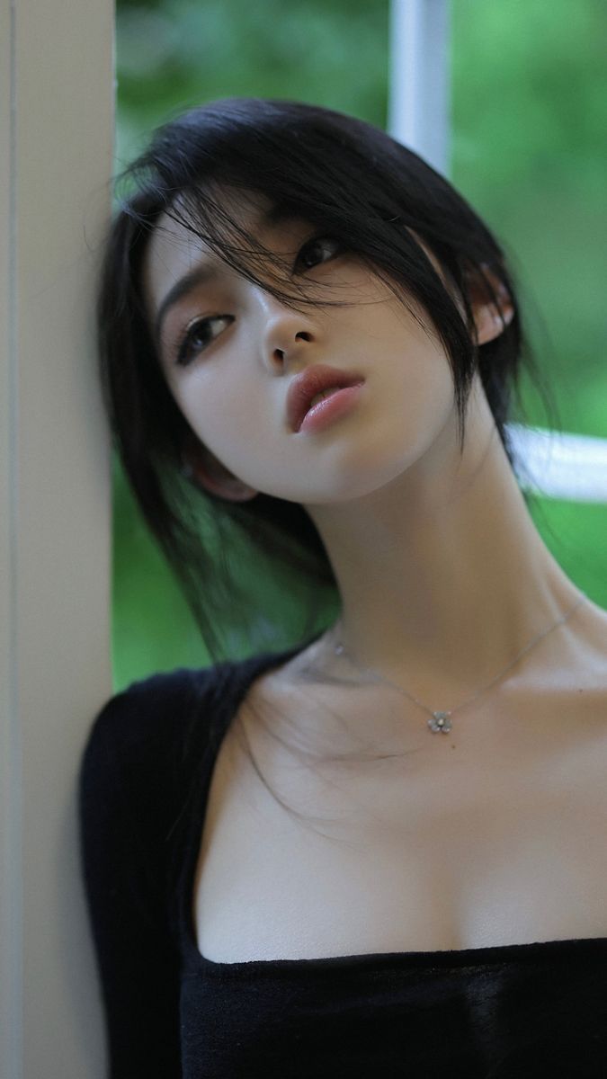 Woman with black hair and a black top, leaning against a wall indoors