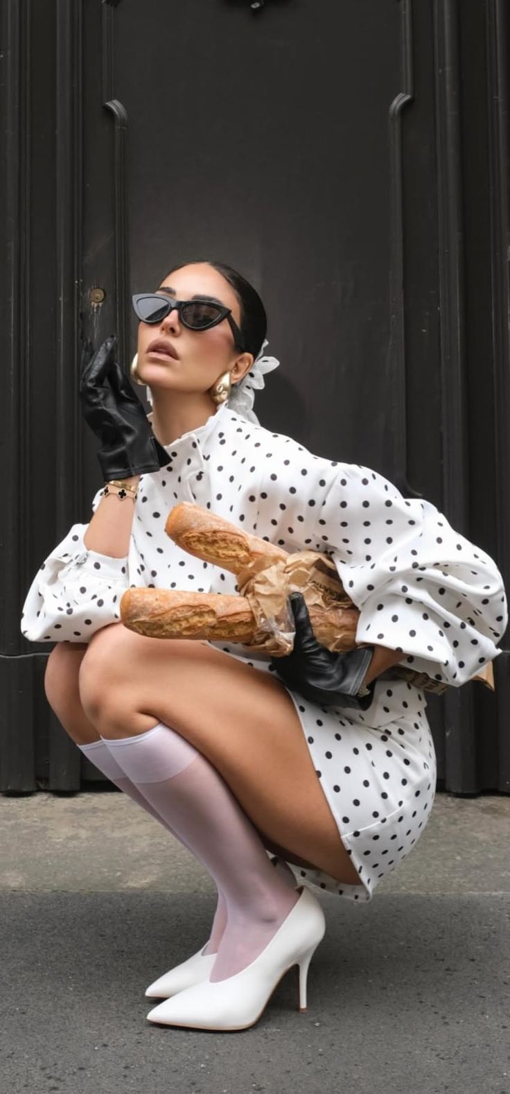 Woman in a polka dot dress and sunglasses holding baguettes in a squatting pose