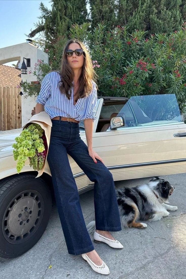 Woman in a striped shirt and jeans with a dog next to a convertible car