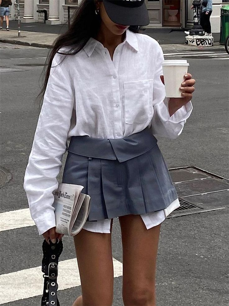 Person in a white shirt and pleated skirt holding coffee and a newspaper on a street