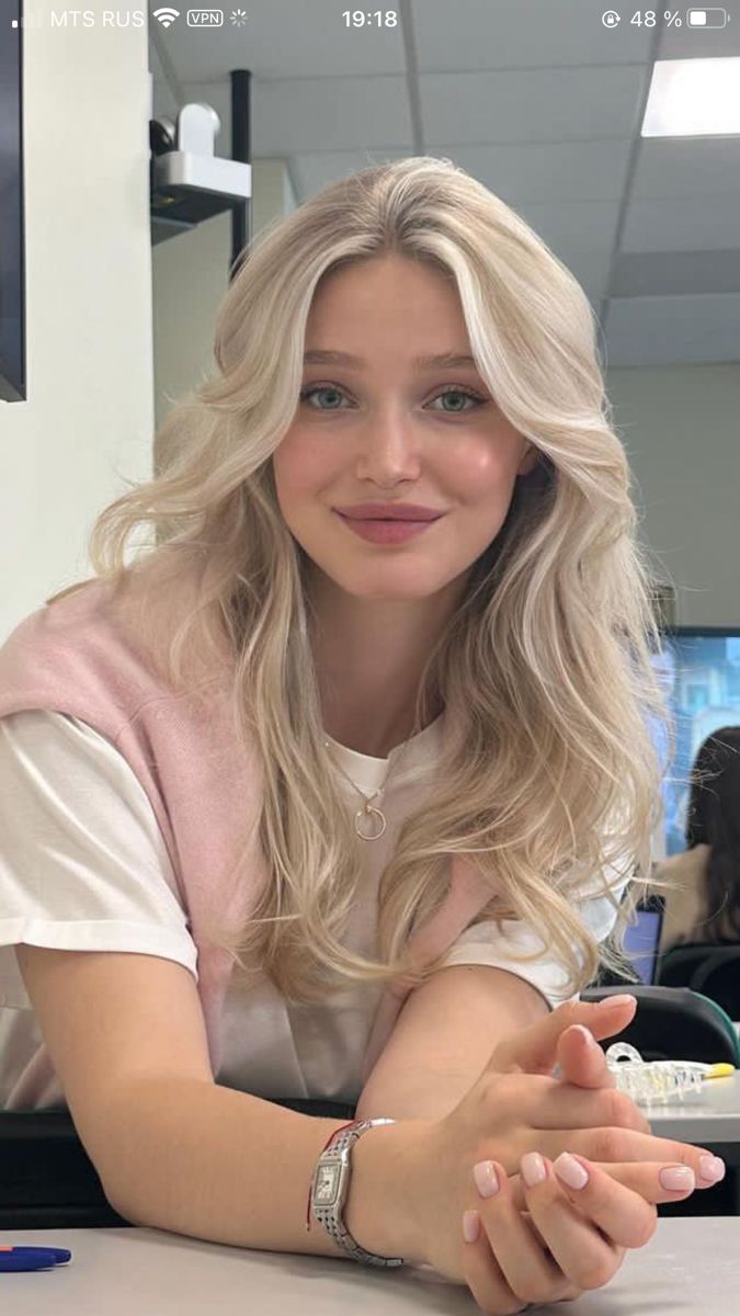 Blonde woman with wavy hair wearing a pink sweater over a white t-shirt indoors
