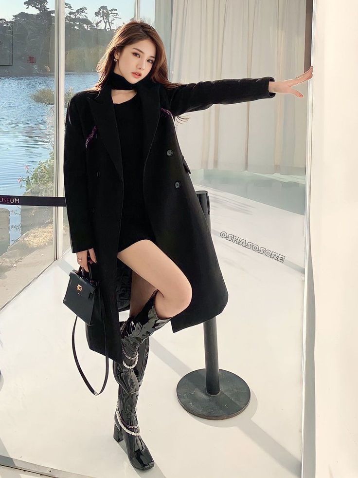 Woman in a black coat and mini dress posing indoors by a window