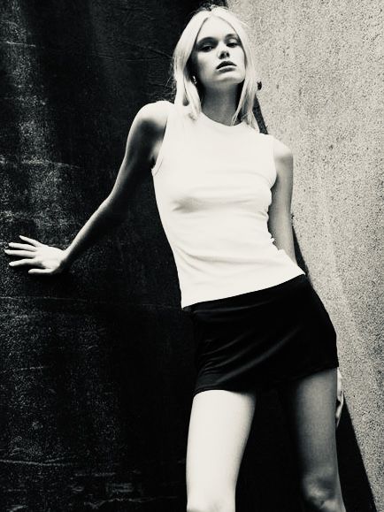 Woman in a white tank top and black mini skirt leaning against a wall