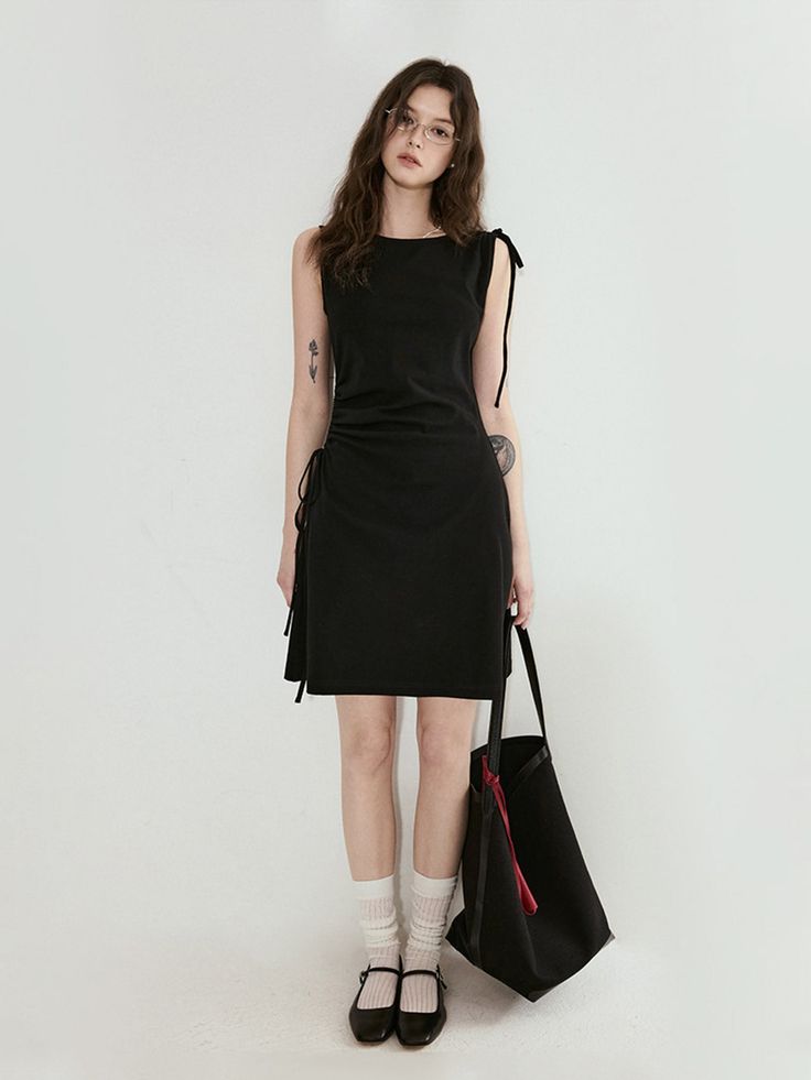 Woman in a black dress and glasses holding a tote bag against a white background