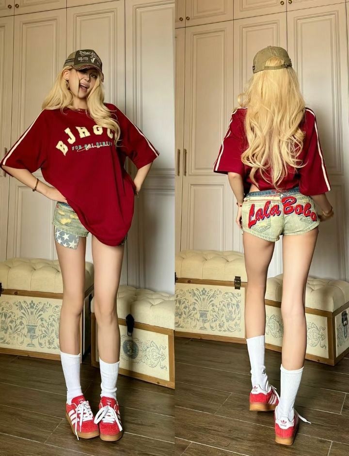 Blonde woman in a red t-shirt, denim shorts, and a baseball cap posing indoors