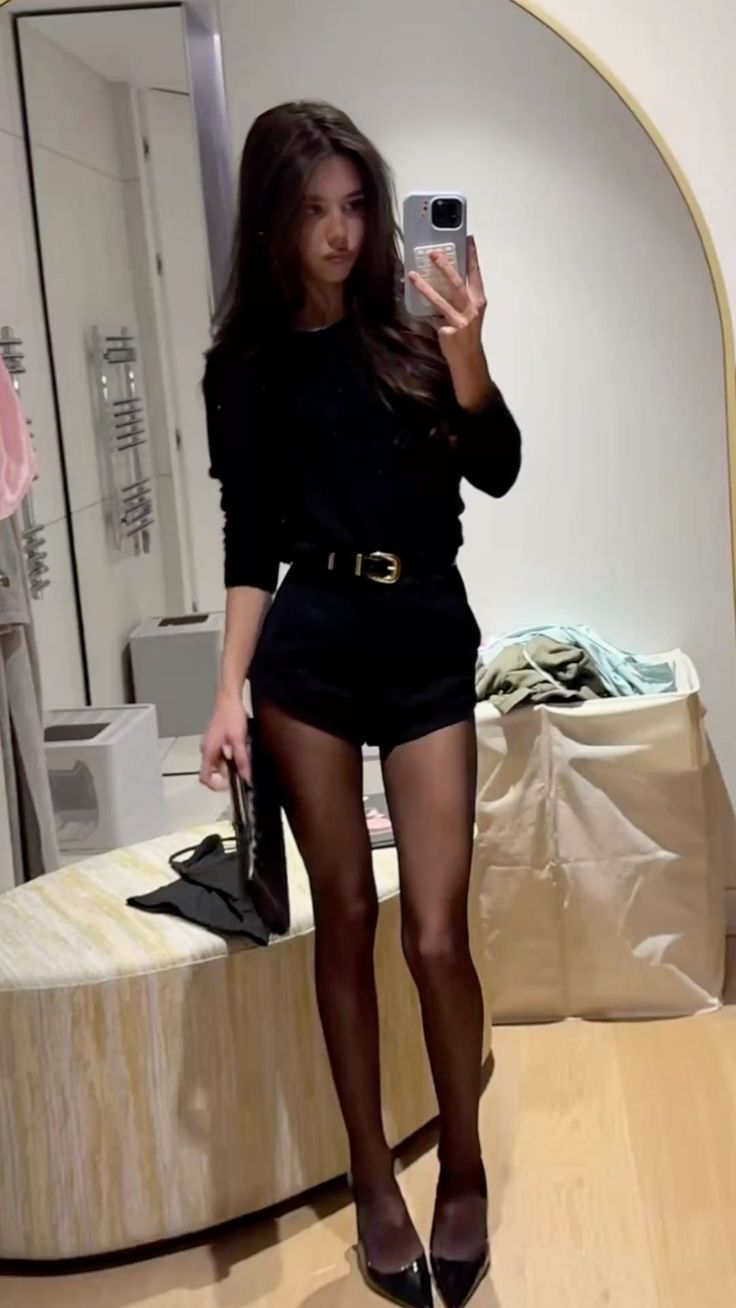 Woman in a black top and shorts with tights taking a mirror selfie