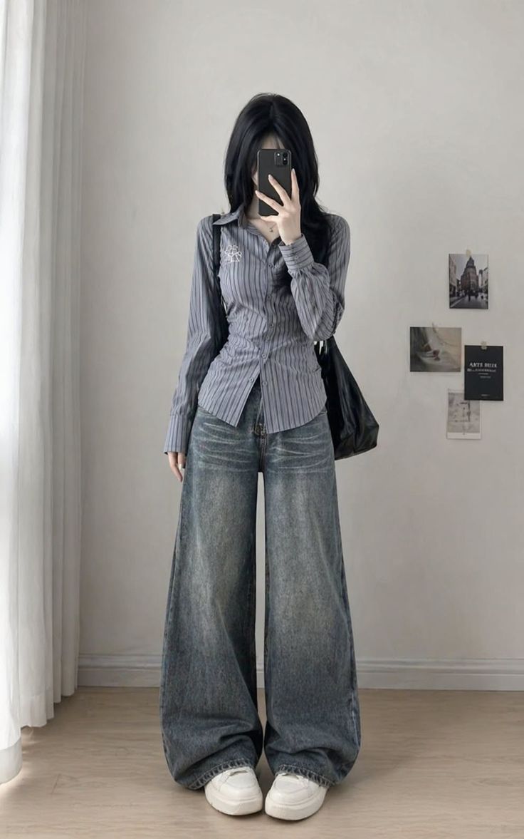 Person with long black hair in a striped shirt and wide leg jeans taking a mirror selfie