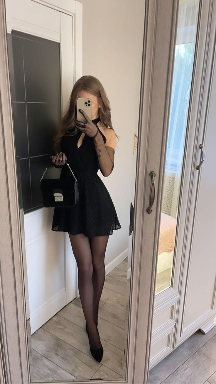 Woman in a black dress and tights taking a mirror selfie indoors