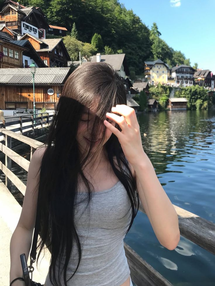 Woman with long dark hair in a grey tank top by a lake in a village