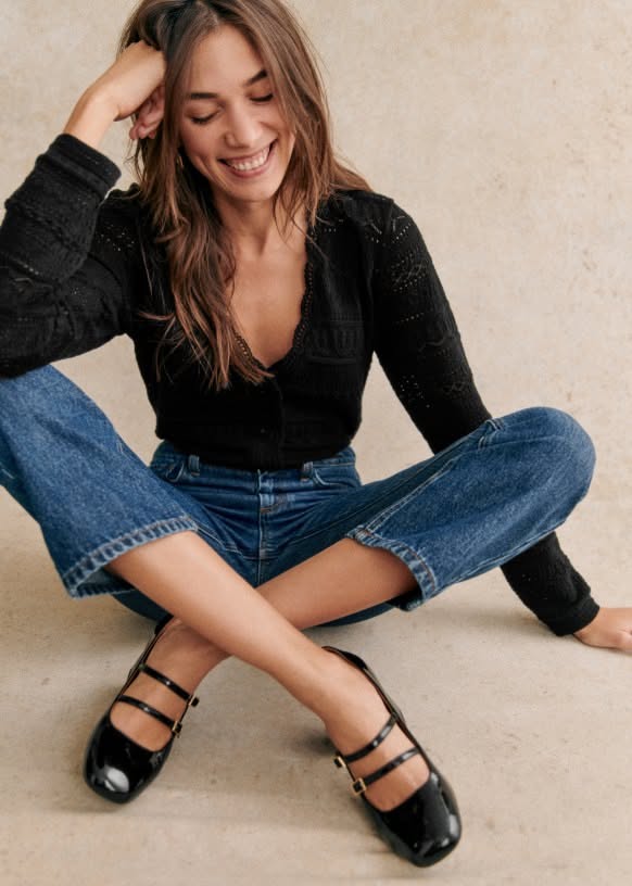 Woman with long hair wearing a black cardigan and blue jeans, sitting and smiling