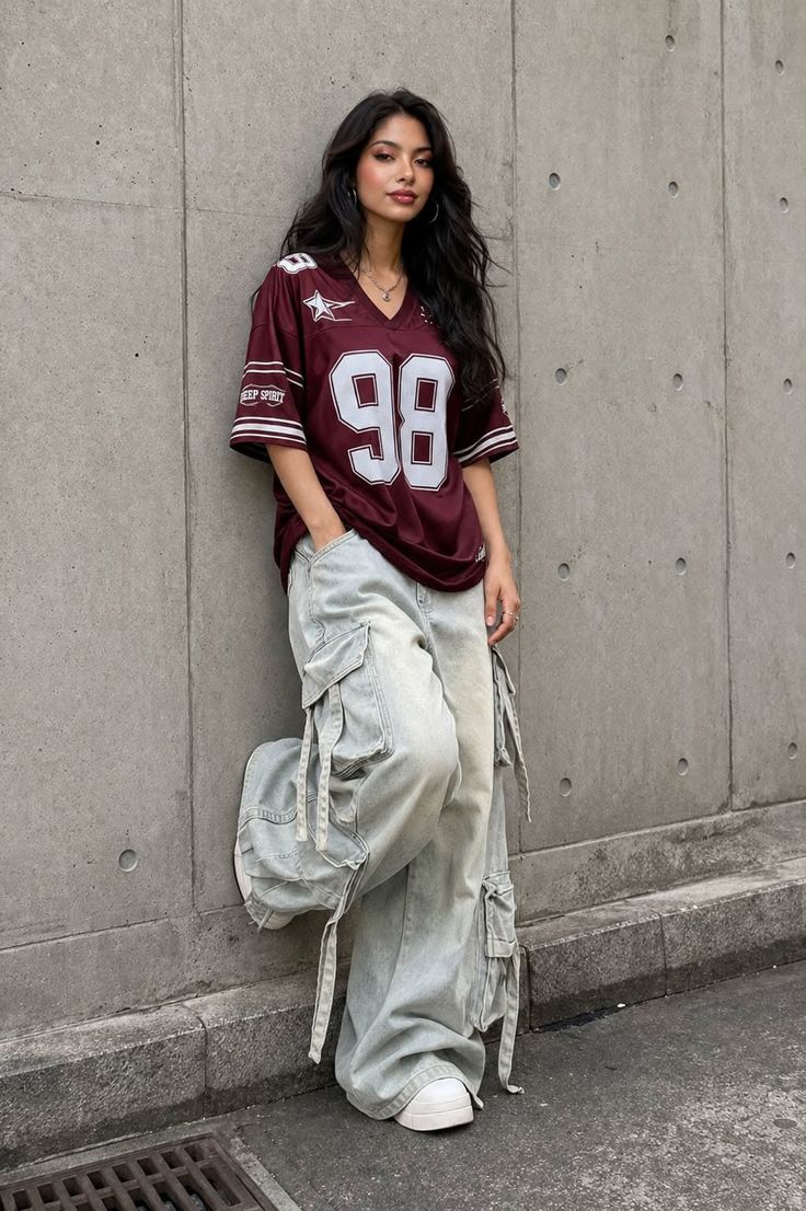 Woman with long dark hair wearing a jersey shirt and cargo pants leaning against a wall