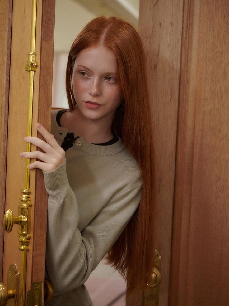 Young woman with long red hair and freckles peeking through a wooden door