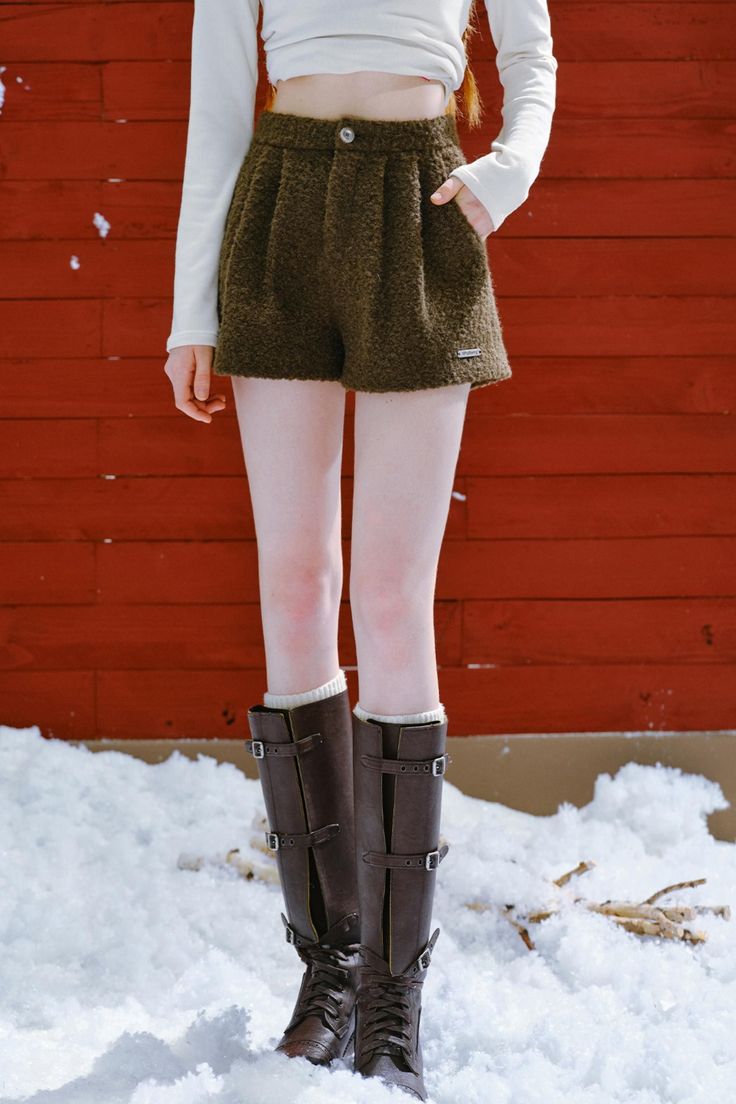 Person in brown shorts and tall boots standing in snow against a red wall
