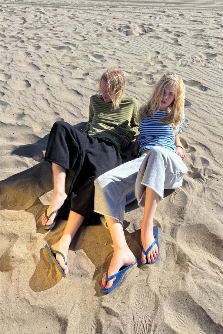 Two people with blonde hair sitting on a sandy beach in flip flops