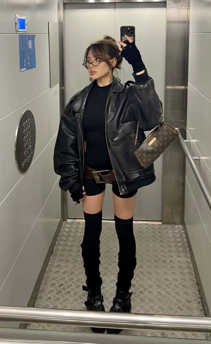 Woman in a black leather jacket and shorts taking a mirror selfie in an elevator