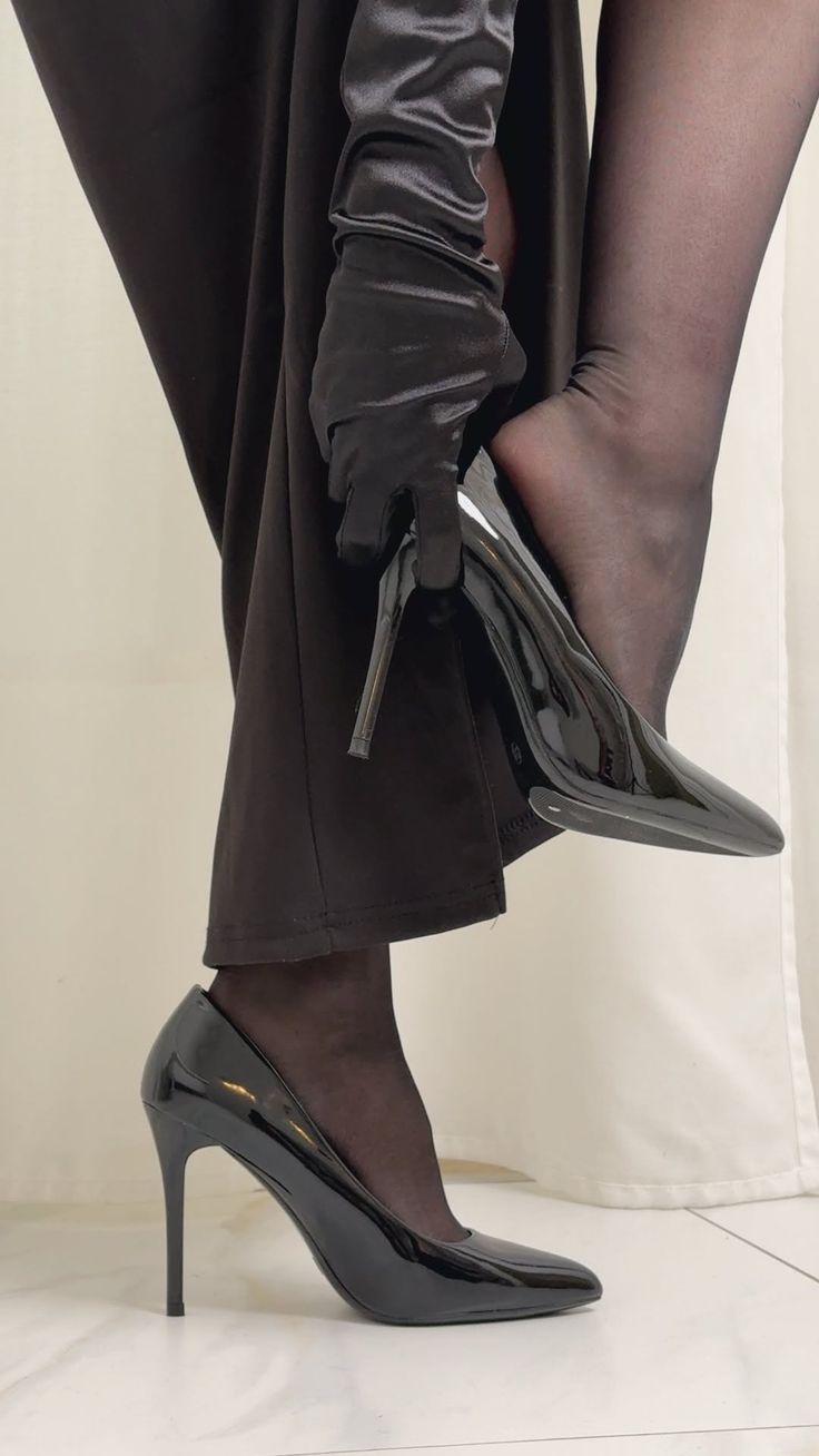 Person in a black dress, gloves, and stockings putting on a black high heel