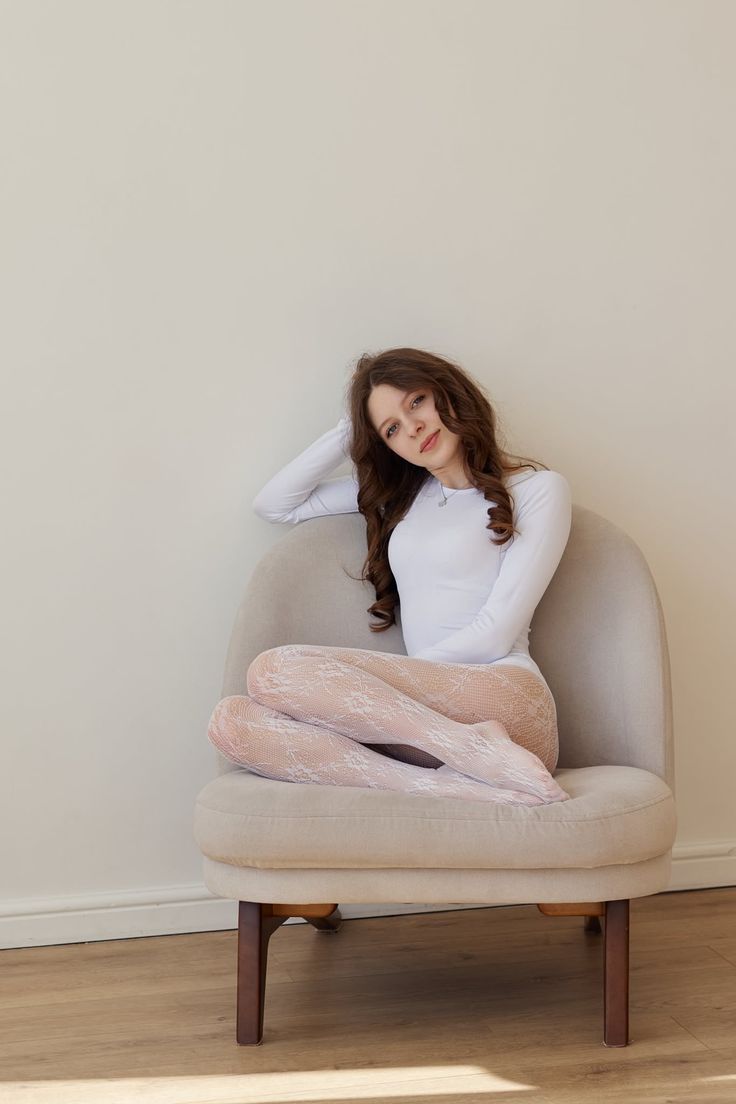 Woman with long curly hair wearing a white bodysuit and lace tights, sitting on a chair