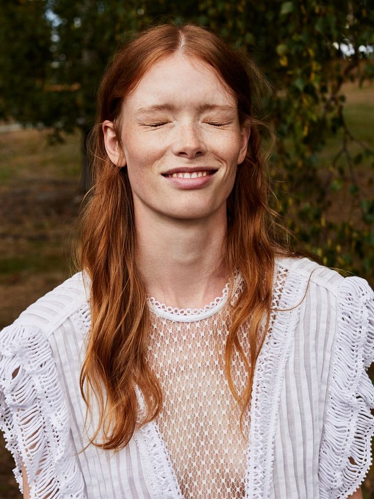 Woman with red hair and freckles smiling with eyes closed in a white lace top