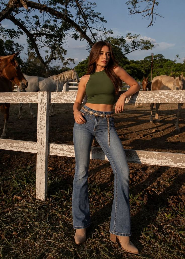 Woman in a crop top and flared jeans leaning on a fence with horses in the background