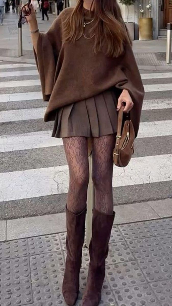 Woman in a brown poncho, pleated skirt, and lace tights standing on a crosswalk