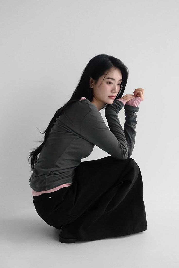 Woman with long black hair wearing a long sleeve shirt and black pants, squatting in a studio