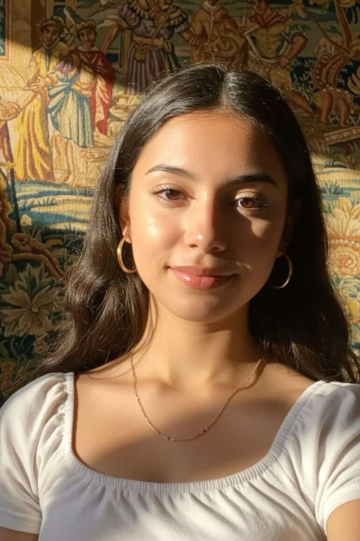 Woman with long dark hair wearing a white top and gold jewelry indoors