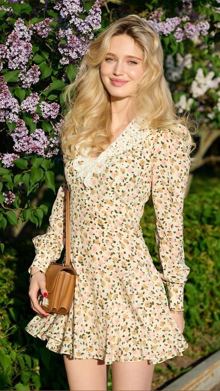 Woman with long blonde hair in a floral dress standing outdoors near lilac flowers